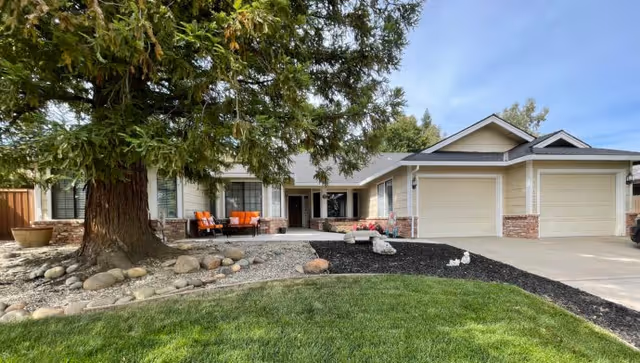 Front exterior view of a single-story house with a large tree and landscaped yard including green grass, rocks, and a driveway leading to a two-car garage. There is outdoor seating with orange cushions near the entrance.