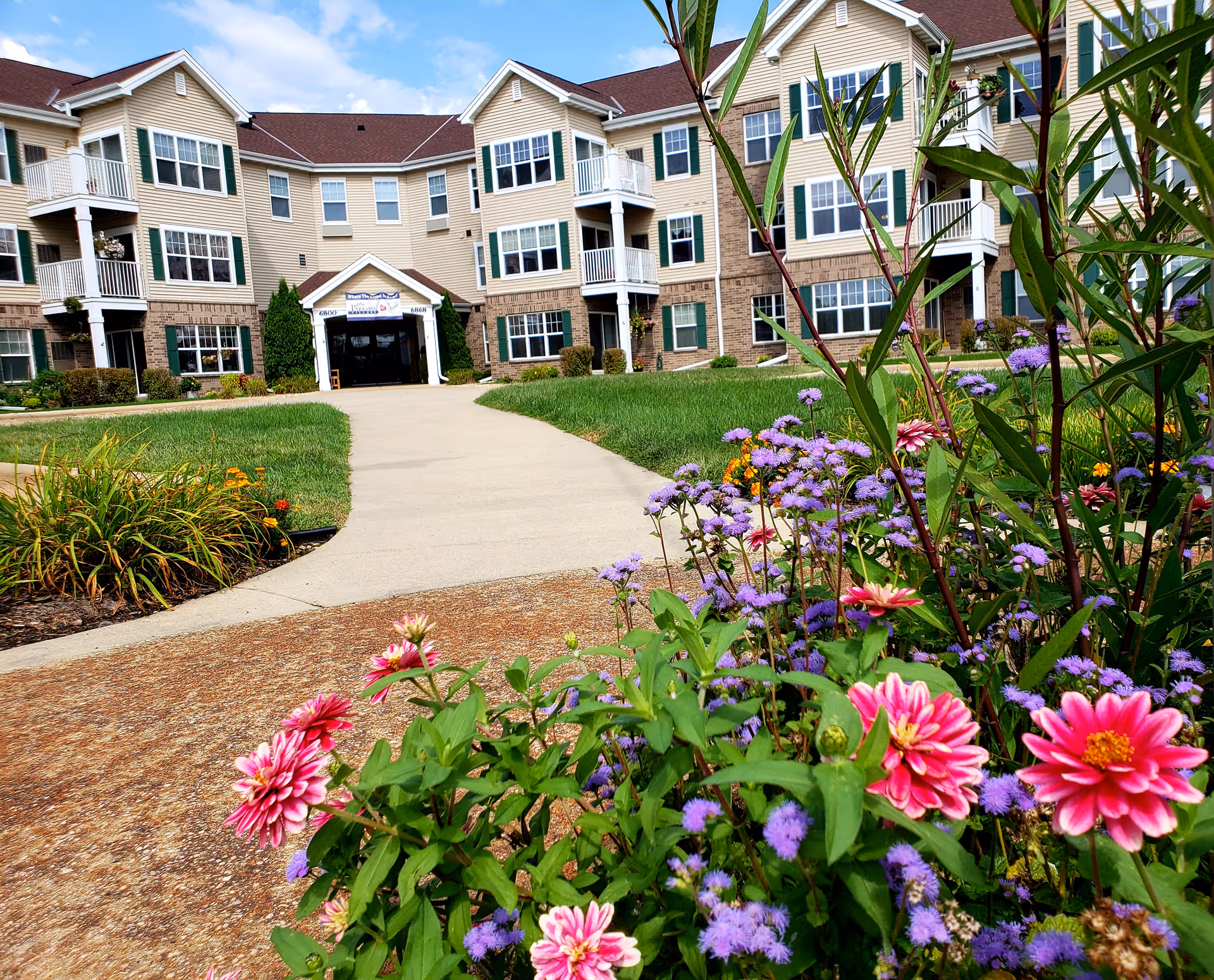 View of the exterior of a multi-story senior living facility named Parkside Village with a paved walkway leading to the entrance. The foreground features colorful flowers and green plants under a blue sky with some clouds.