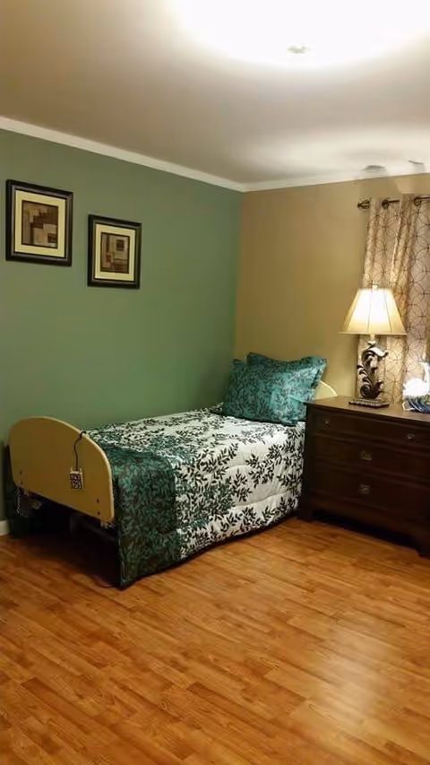 A small bedroom with a single bed covered in a green and white floral bedspread and matching pillows. The bed has a light wooden headboard and footboard. Next to the bed is a dark wooden nightstand with a decorative lamp and a curtain with a circular pattern hanging on the window. The walls are painted green and beige, and the floor is wooden.