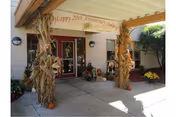 Entrance area of a facility decorated for a 20th anniversary celebration with a banner reading 'Happy 20th Anniversary Oakleaf'. The entrance has double glass doors framed in red, with two columns wrapped in dried cornstalks and pumpkins placed around the base. There are potted plants and flowers near the entrance under a covered walkway.