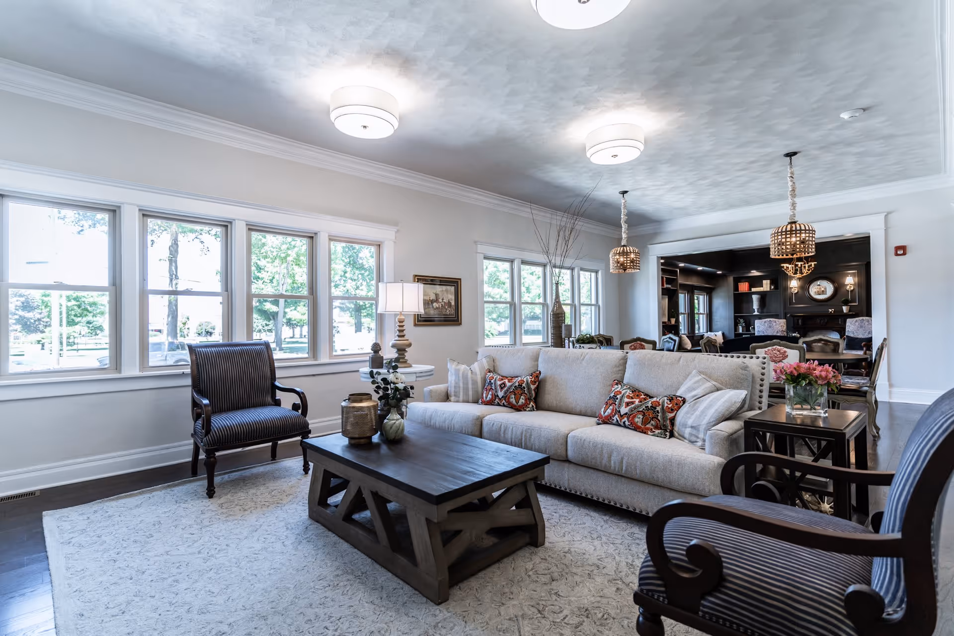 A bright and spacious living room with large windows letting in natural light. The room features a beige sofa with patterned cushions, two dark wooden armchairs with striped upholstery, a wooden coffee table with decorative items, and a side table with a lamp and flowers. In the background, there is a dining area with a dark wood table and chairs, pendant lights, and built-in shelves with decor.