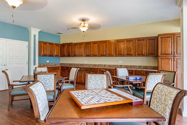 A well-lit room with wooden cabinets lining the back wall and several tables with cushioned chairs arranged around the room. One table in the foreground has a Scrabble board game set up on it. The walls are painted in light yellow and teal colors, and the floor is wooden.