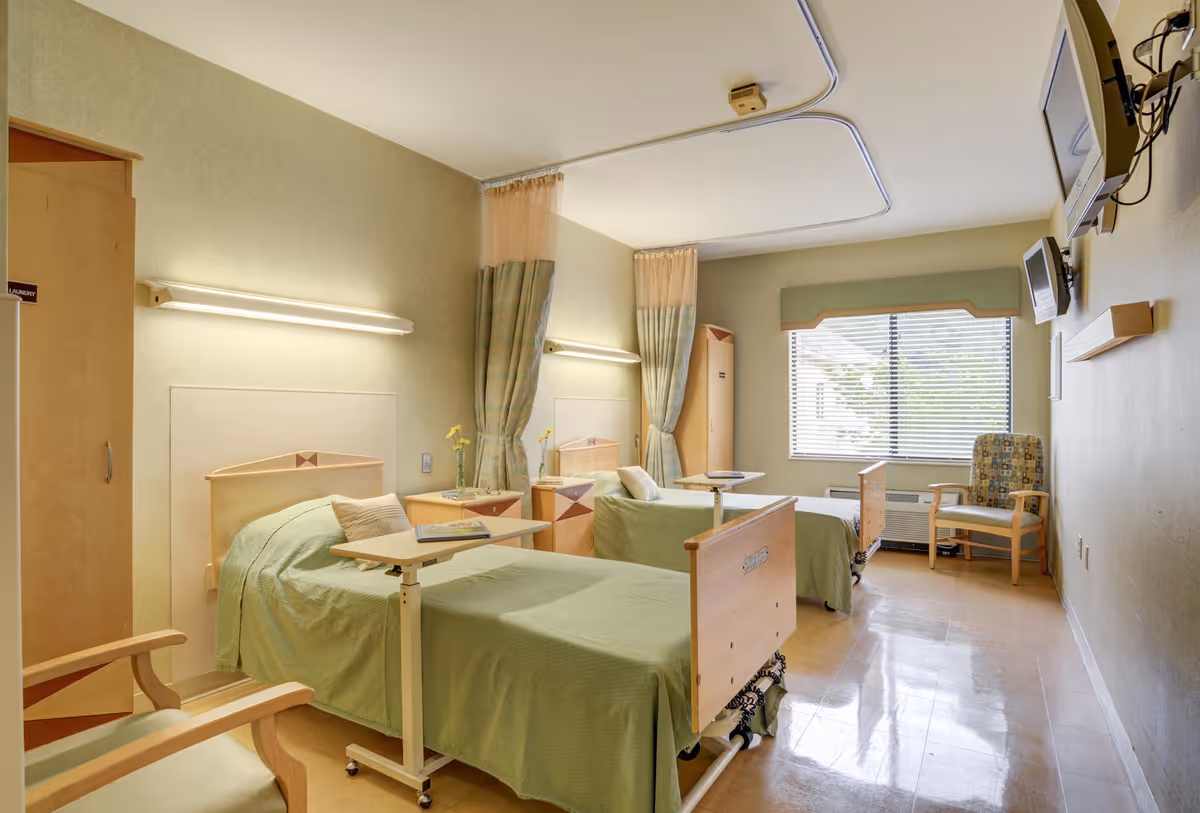 A bright and clean room with two hospital-style beds covered in green bedspreads, each with an adjustable overbed table. The room has light green walls, two wall-mounted lights above the beds, a window with blinds and a green valance, a patterned armchair, and two wall-mounted televisions. There are privacy curtains between the beds and a wooden cabinet labeled 'Laundry' in the corner.
