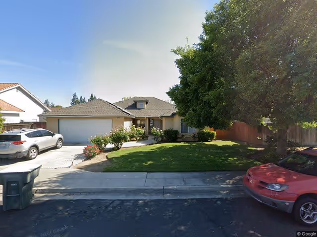 Single-story house with a two-car garage, a well-maintained front lawn with bushes and trees, a driveway with a silver SUV parked, and a red car parked on the street in front of the house under a clear sky.