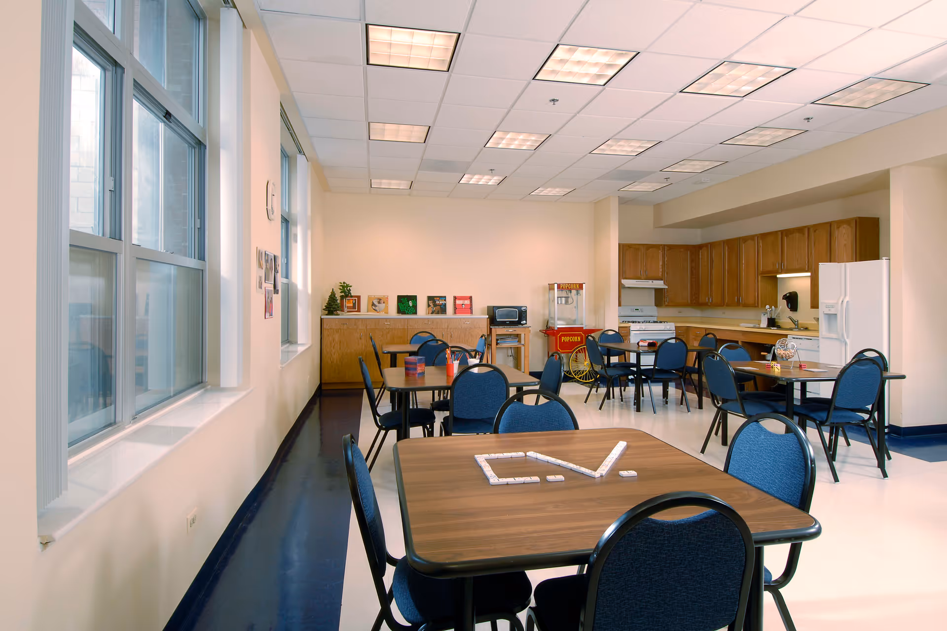 A bright communal room with several tables and blue chairs arranged for group activities. The room features large windows on one side, a kitchenette with wooden cabinets, a refrigerator, stove, and a popcorn machine. On the tables are game pieces and supplies, suggesting a recreational or dining area.