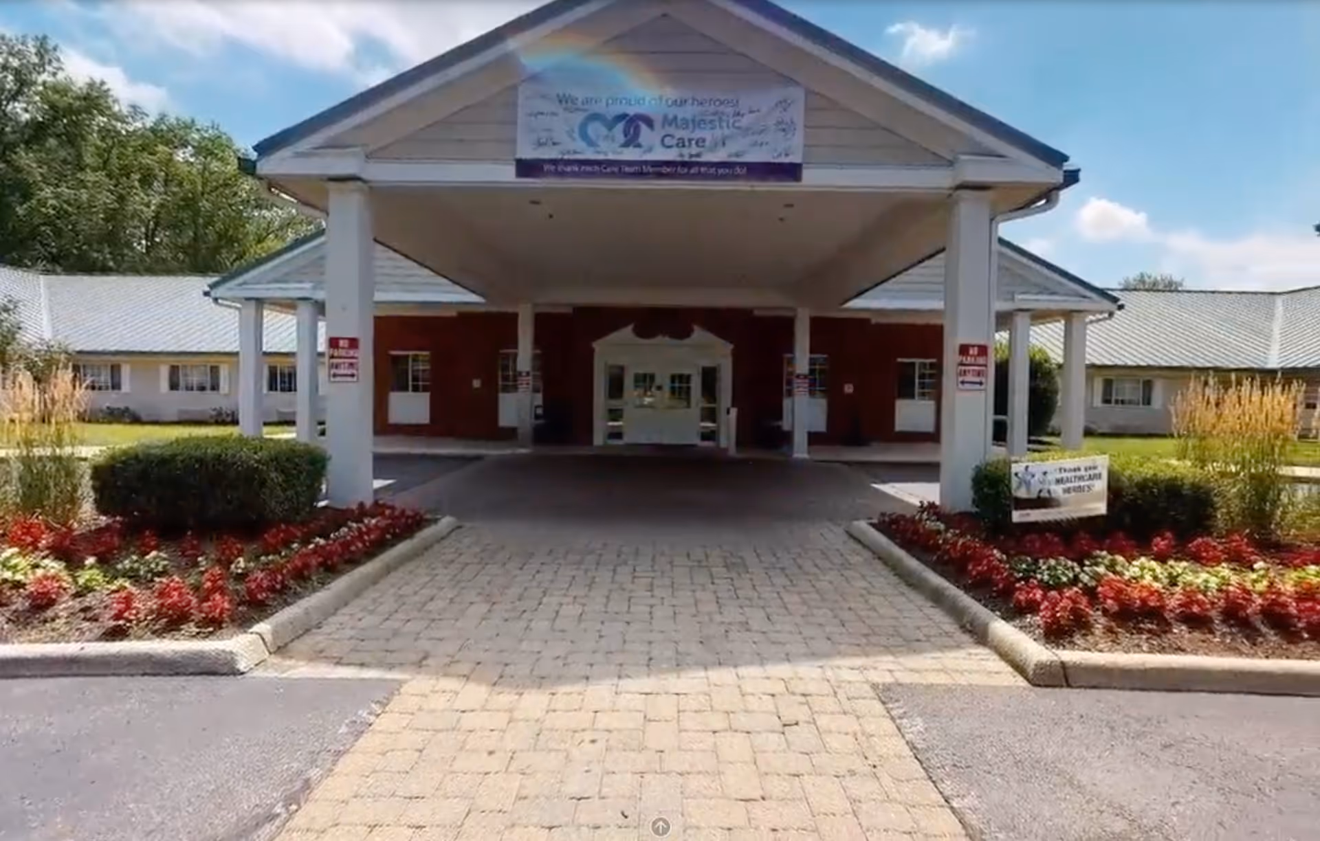 Front entrance of Majestic Care of Whitehall facility with a covered driveway, landscaped flower beds on both sides, and a banner above the entrance that reads 'We are proud of our heroes! Majestic Care'.