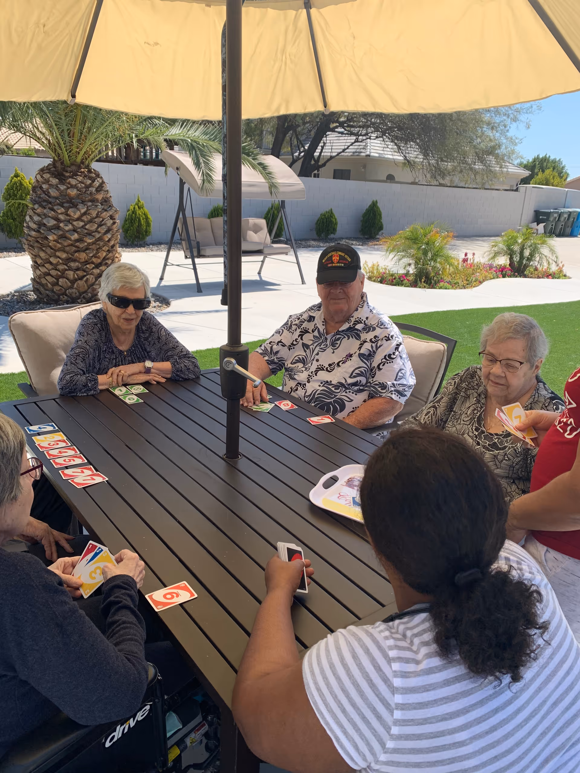 A group of elderly people sitting around a rectangular outdoor table under a large beige umbrella, playing a card game. The setting is a sunny outdoor patio area with palm trees, green artificial grass, and a swing bench in the background.