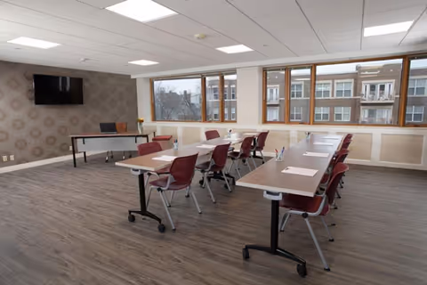 A bright meeting or conference room with two long tables arranged in a U-shape, surrounded by maroon chairs. The room has large windows letting in natural light and a wall-mounted flat screen TV. The floor is covered with wood-patterned vinyl, and the walls are light-colored with a patterned accent wall behind the TV.