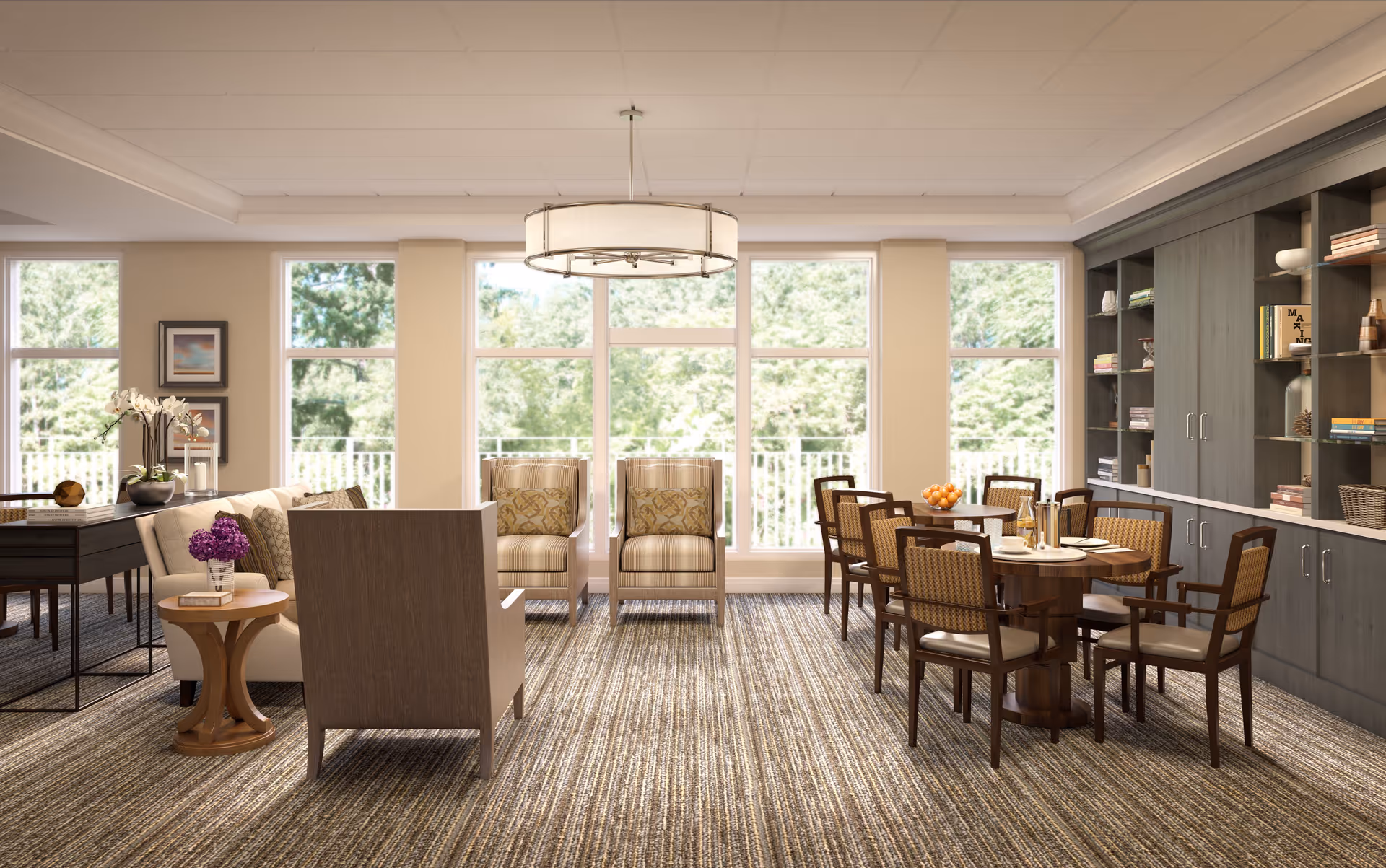 A bright and spacious senior living common area with large windows letting in natural light. The room features comfortable seating including armchairs and a sofa, a round dining table with chairs, a side table with a vase of purple flowers, and built-in shelving filled with books and decorative items. The carpet has a striped pattern and a modern chandelier hangs from the ceiling.