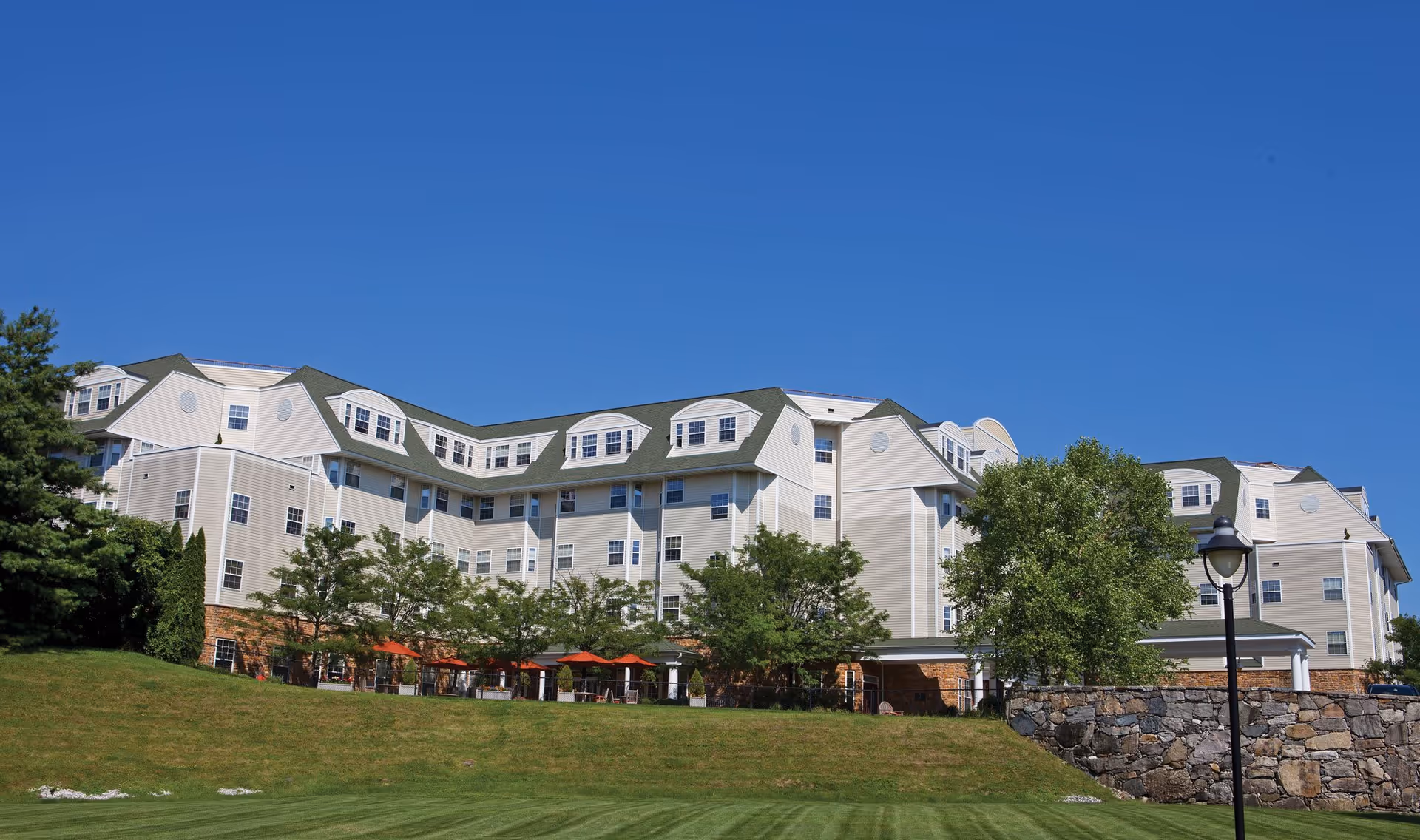 Exterior view of a large, multi-story senior living facility building with beige siding and green roofs, surrounded by trees and a well-maintained grassy lawn under a clear blue sky.