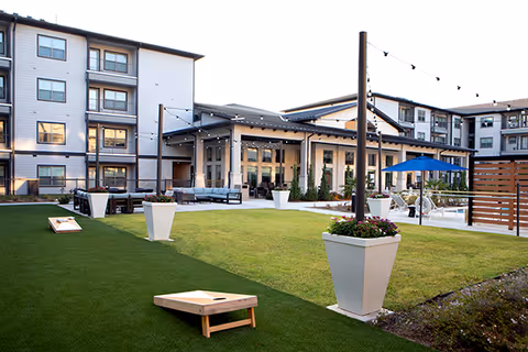 Outdoor courtyard area of a senior living facility with green lawn, cornhole game boards, large white planters with flowers, string lights overhead, seating areas with couches and chairs, and multi-story residential buildings in the background.