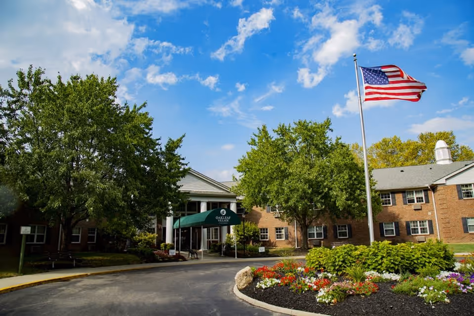 Exterior view of Oakleaf Village of Columbus senior living facility with a circular driveway, landscaped flower beds, green trees, and an American flag flying on a flagpole under a partly cloudy blue sky.