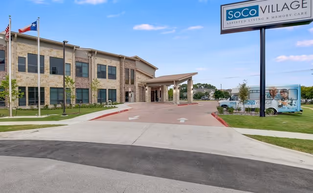 Exterior view of SoCo Village assisted living and memory care facility showing a two-story building with stone and brick facade, a covered entrance driveway, three flagpoles with flags, a large sign with the facility name, and a branded van parked on the grass.