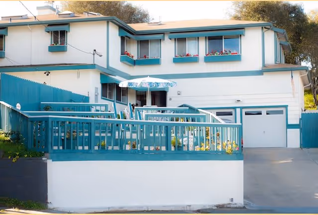 Exterior view of a two-story senior living facility with white walls and teal trim. The building features multiple windows with flower boxes, a teal railing around a patio area with outdoor furniture and an umbrella, and a driveway leading to a garage.
