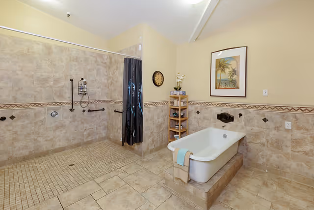 A spacious bathroom with beige tiled walls and floor. There is a walk-in shower area with a black curtain and grab bars, and a white bathtub with towels draped over the side. A wooden shelving unit holds toiletries, and a framed picture of a beach scene hangs on the wall above the bathtub. A round clock is mounted on the wall near the shower.