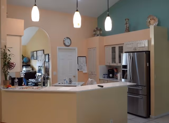Interior view of a kitchen area with a beige and green color scheme. The kitchen features a stainless steel refrigerator, white cabinets, a countertop with a sink, and three hanging pendant lights. There is an archway leading to another room with office equipment and wall decorations visible.