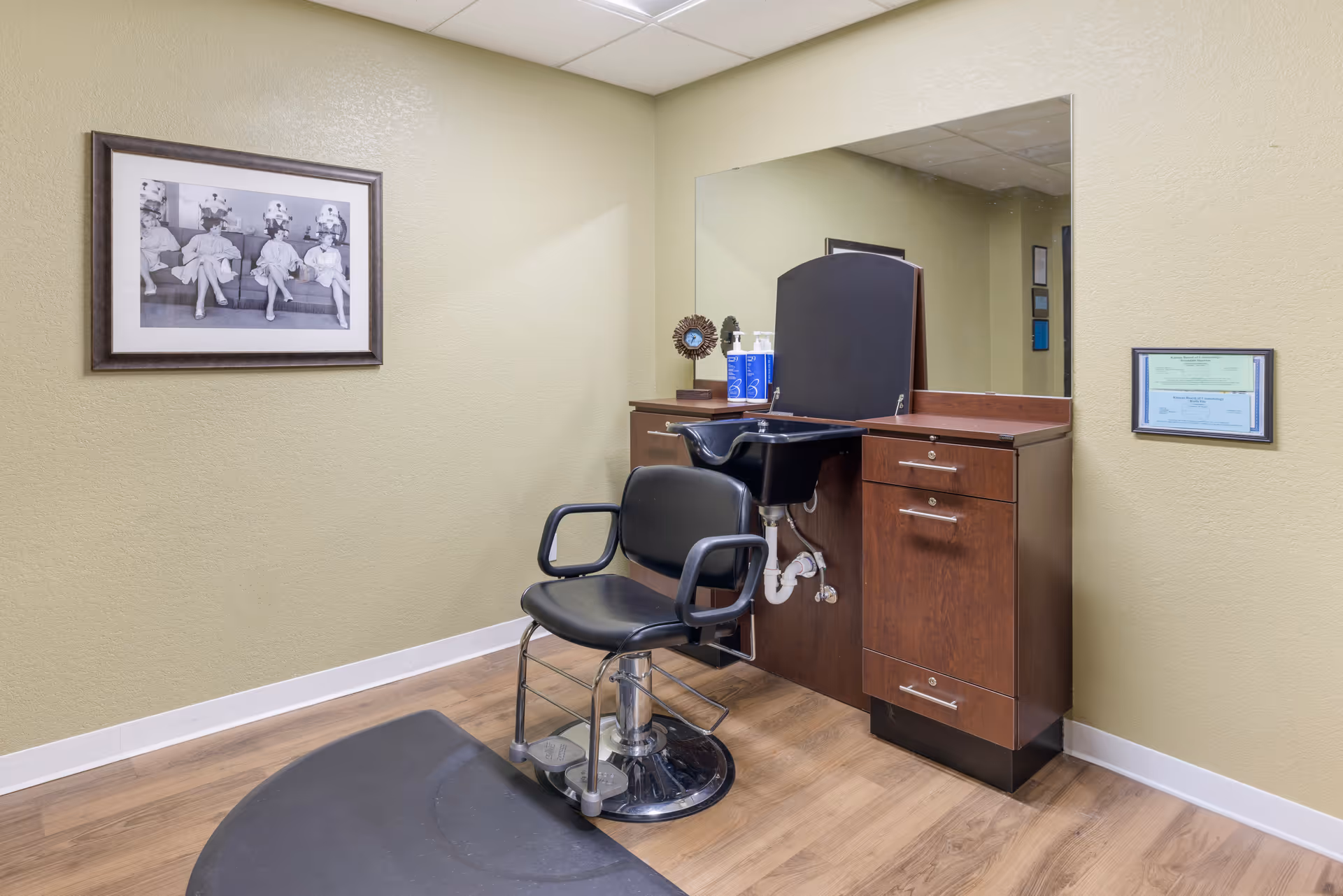 A small salon area with a black salon chair in front of a wooden cabinet with a built-in black sink. A large mirror is mounted on the wall above the cabinet. The walls are light green, and there is a framed black and white photo of four women sitting on a couch with hair dryers on their heads on the left wall. A framed certificate is hung on the right wall. The floor is wood with a black mat in front of the chair.
