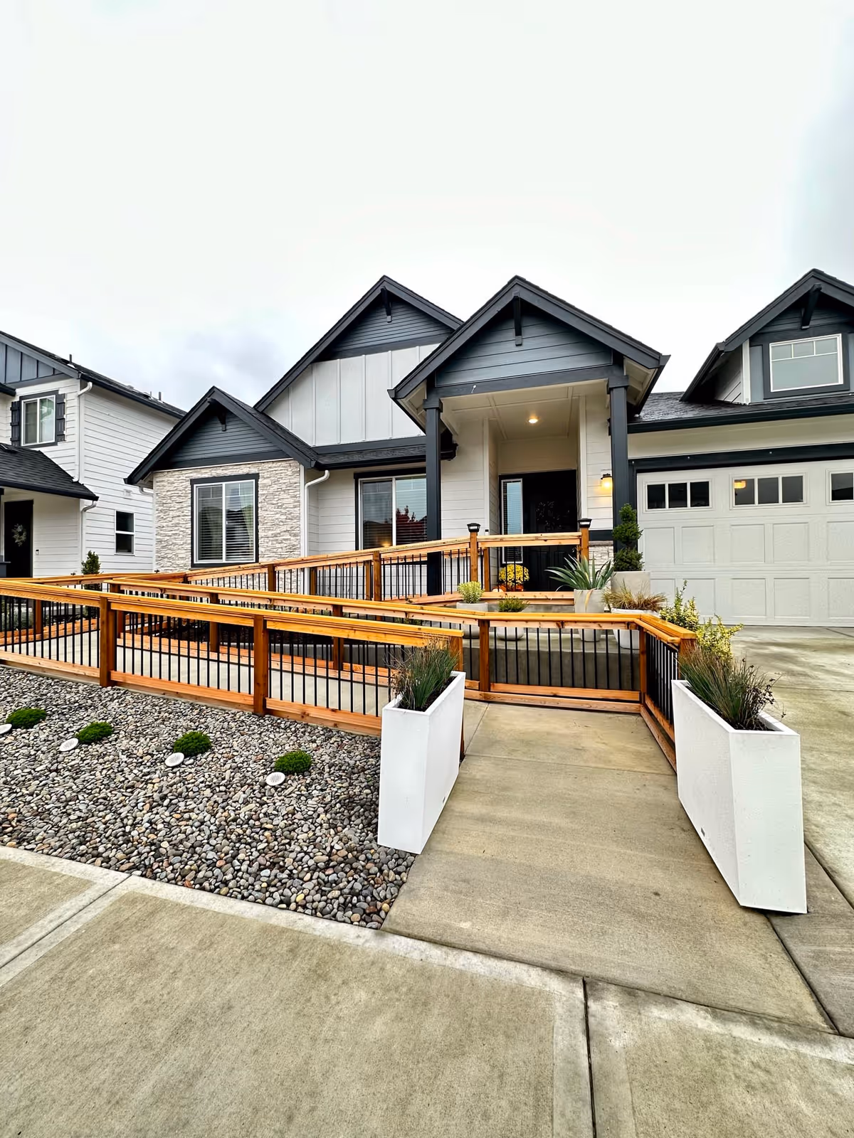 Front exterior view of a modern single-story house with a gray and white color scheme, featuring a wooden ramp with black railings leading to the front door, two white planters with plants on either side of the ramp entrance, and a garage on the right side.