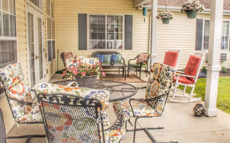 A covered outdoor patio area with floral cushioned metal chairs around a round metal table, a flower pot centerpiece, two red cushioned rocking chairs, and a bench with pillows against the wall of a yellow building with windows and white trim.