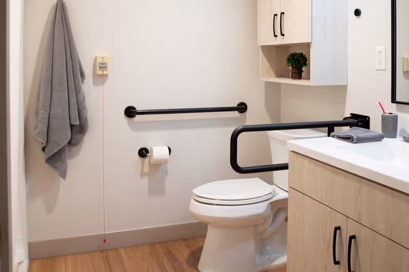 A clean and accessible bathroom with a white toilet equipped with black grab bars on the side and behind it. There is a toilet paper holder mounted on the wall, a gray towel hanging on a hook, and a wooden vanity with a white countertop. On the countertop, there is a folded gray towel and a gray toothbrush holder with a pink toothbrush. Light wood cabinets are mounted above the toilet.