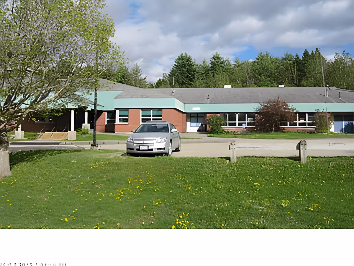 Exterior view of Frankfort Assisted Living facility showing a single-story building with a gray roof and teal trim. There is a silver car parked in front of the building on a paved area, surrounded by green grass with yellow flowers and trees. The sky is partly cloudy.