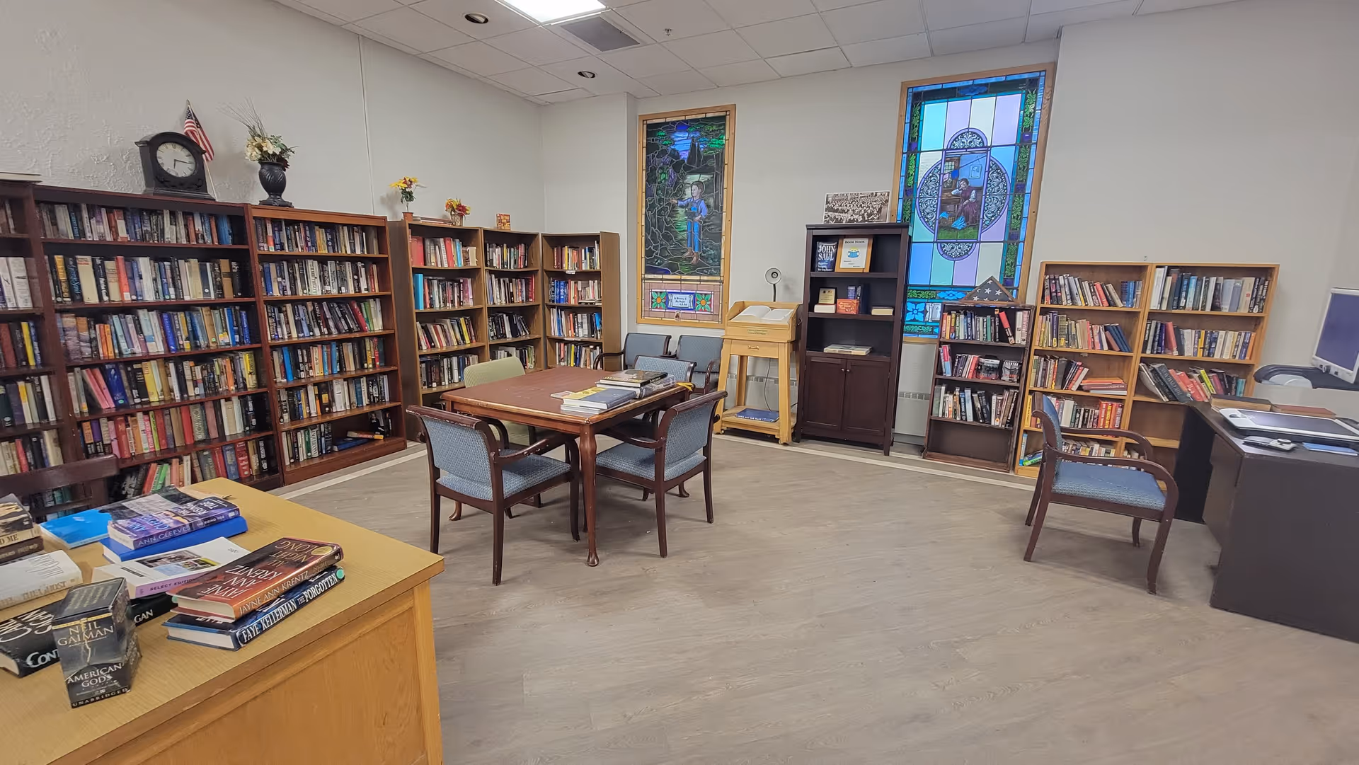 A well-lit library room with multiple wooden bookshelves filled with books, a wooden table with four chairs in the center, stained glass windows on the far wall, and a desk with a computer on the right side.