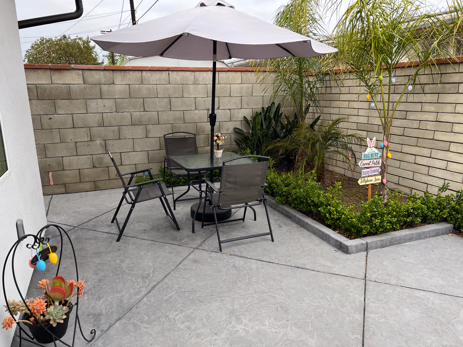Outdoor patio area with a glass table and four chairs under a large white umbrella. The patio is surrounded by a low garden bed with green plants and small palm trees. A decorative sign with Easter-themed directions is placed in the garden bed. The area is enclosed by a beige brick wall.
