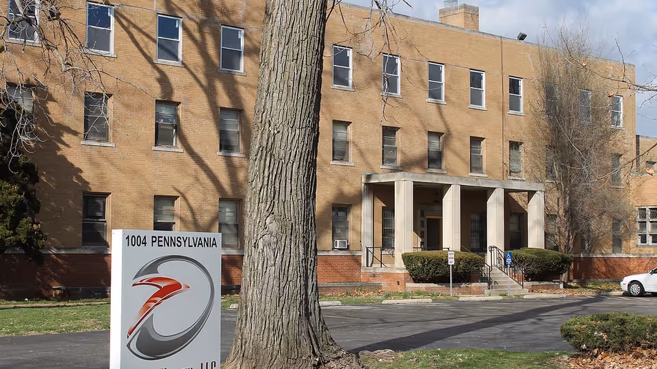 Exterior view of a three-story brick building with multiple windows and a small entrance porch with columns. In front of the building is a large tree and a sign that reads '1004 PENNSYLVANIA' with a logo. There is a parking lot with a white car visible on the right side.