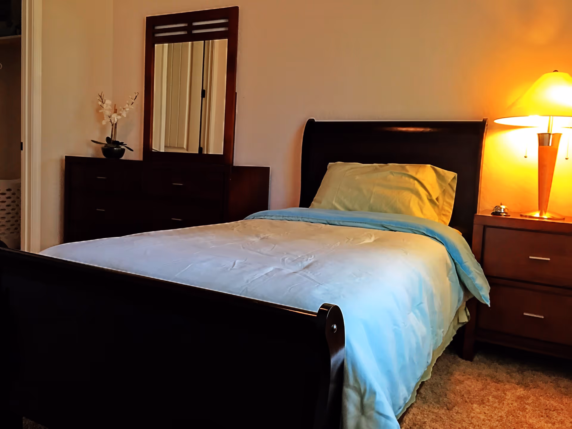 A cozy bedroom with a single bed made up with light blue and beige bedding. Next to the bed is a wooden nightstand with a lit lamp and a small bell. Across from the bed is a dark wooden dresser with a mirror and a small vase with white flowers. The room has beige walls and carpeted flooring.