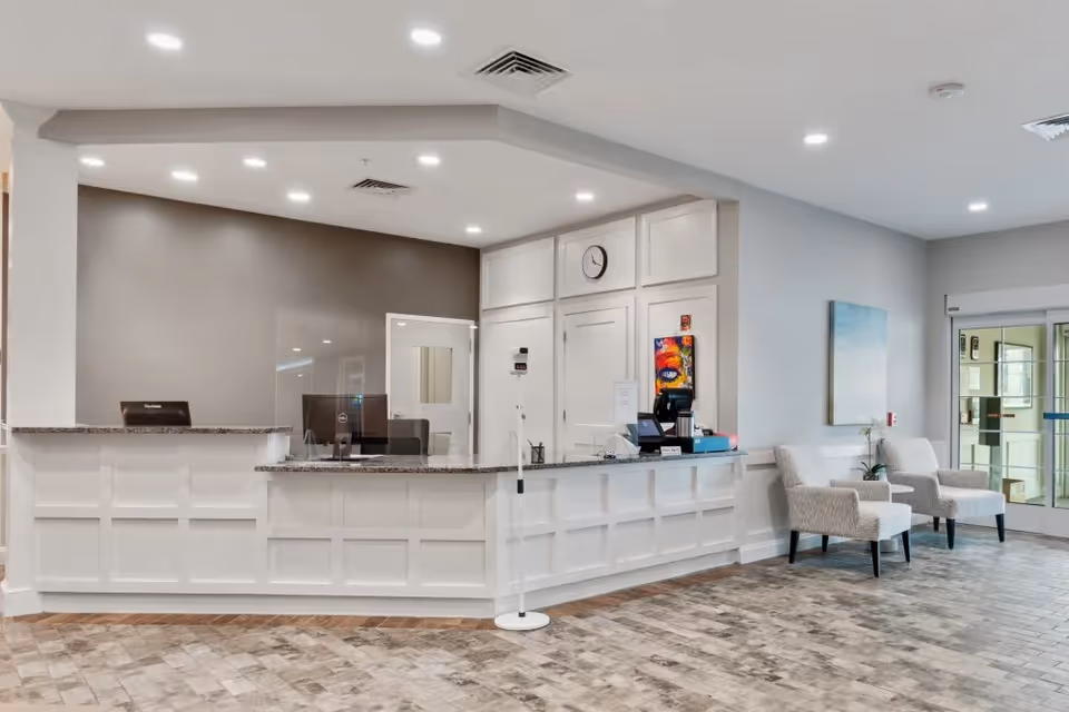 Reception area of Merrill Gardens at West Chester featuring a white paneled front desk with granite countertop, two computer monitors, a clock on the wall behind, and two upholstered chairs with a small table and plant near the entrance door.