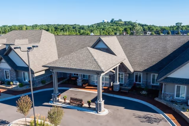 Front exterior view of a single-story senior living facility with a covered entrance, stone and siding facade, surrounded by a paved driveway and greenery in the background under a clear blue sky.