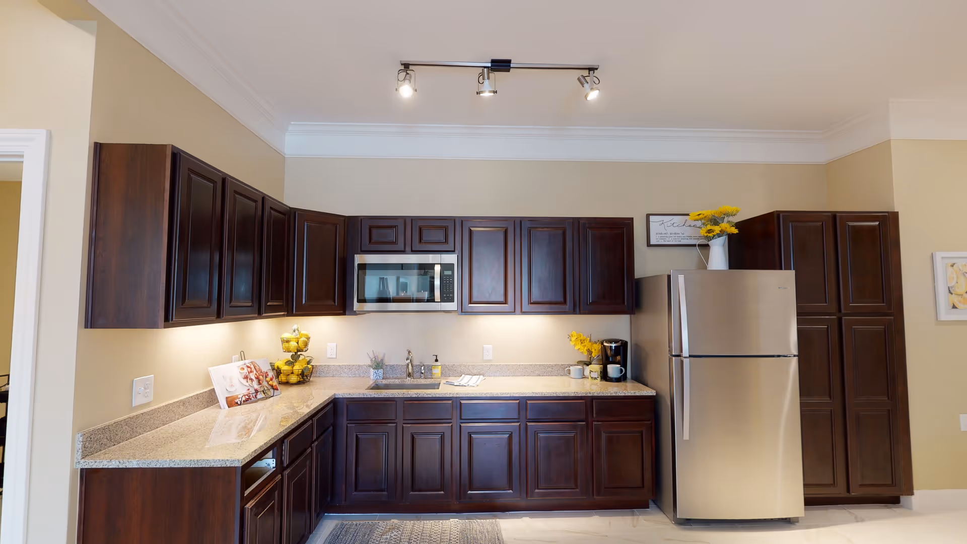 A modern kitchen with dark wood cabinets, granite countertops, a stainless steel refrigerator, and a built-in microwave. The countertop has a tiered fruit basket with lemons, a small plant, a coffee maker with two mugs, and a decorative sign with yellow flowers on top of the refrigerator. The kitchen is well-lit with under-cabinet lighting and ceiling track lights.