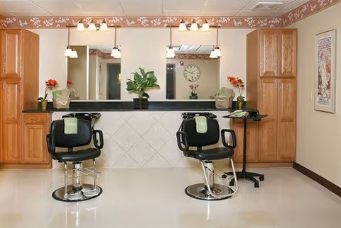 Interior view of a salon area with two black salon chairs facing a counter with a large mirror. The counter has plants and towels on it, with wooden cabinets on either side. The walls are beige with a floral border near the ceiling, and there is a clock and framed artwork visible in the background.