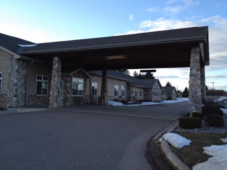 Exterior view of Lakewood Assisted Living facility showing a covered entrance supported by stone pillars, with a driveway leading up to the building. Snow is visible on the ground and bushes are planted near the curb.