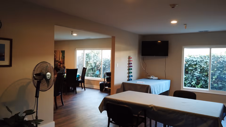 Interior view of a senior living facility room with tables covered in light blue tablecloths, chairs around the tables, a standing fan, a wall-mounted TV, and large windows showing greenery outside. The room has wooden flooring and neutral-colored walls.