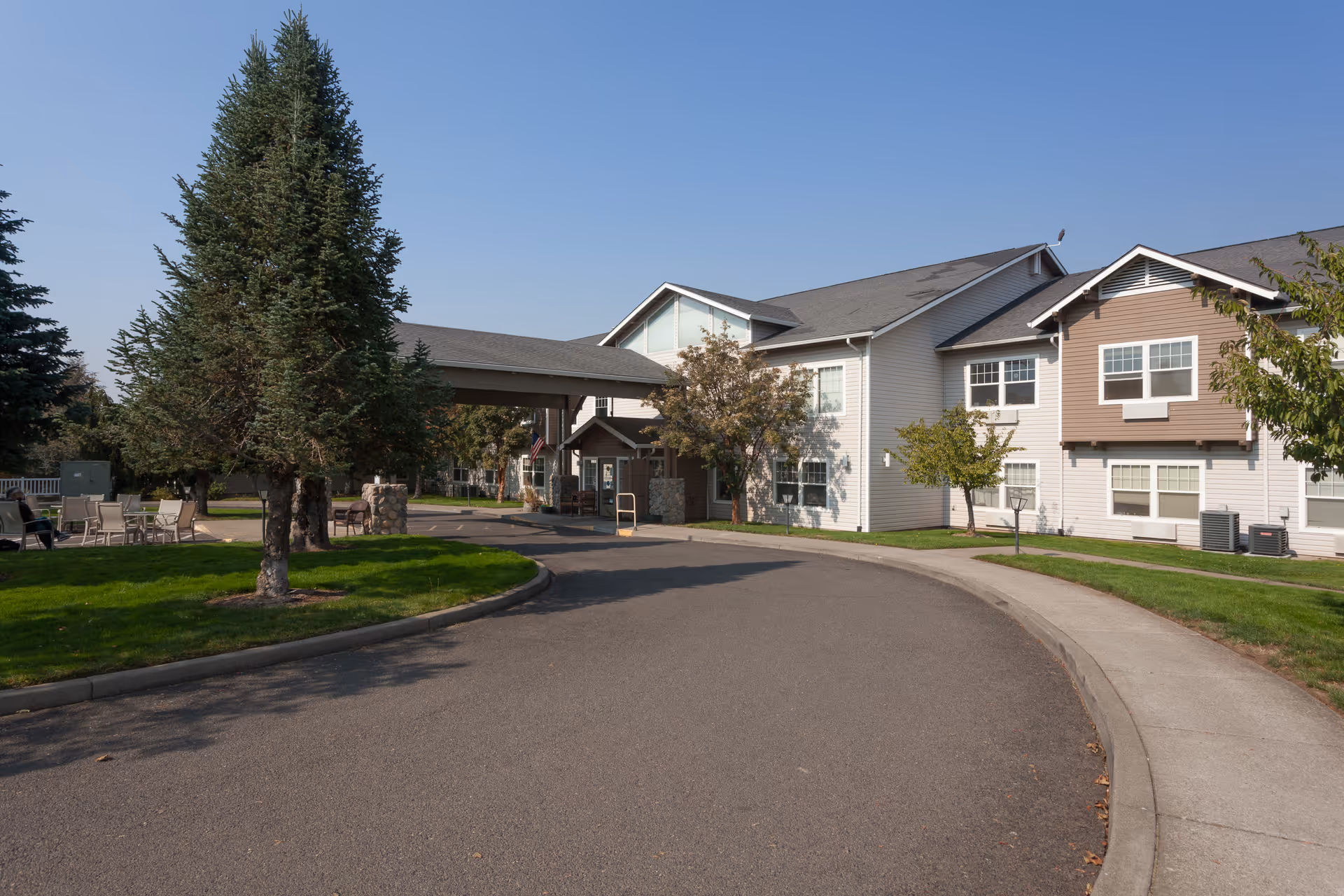 Exterior view of Skylark Assisted Living facility showing a two-story building with a covered entrance, surrounded by trees, green lawns, and a paved driveway under a clear blue sky.