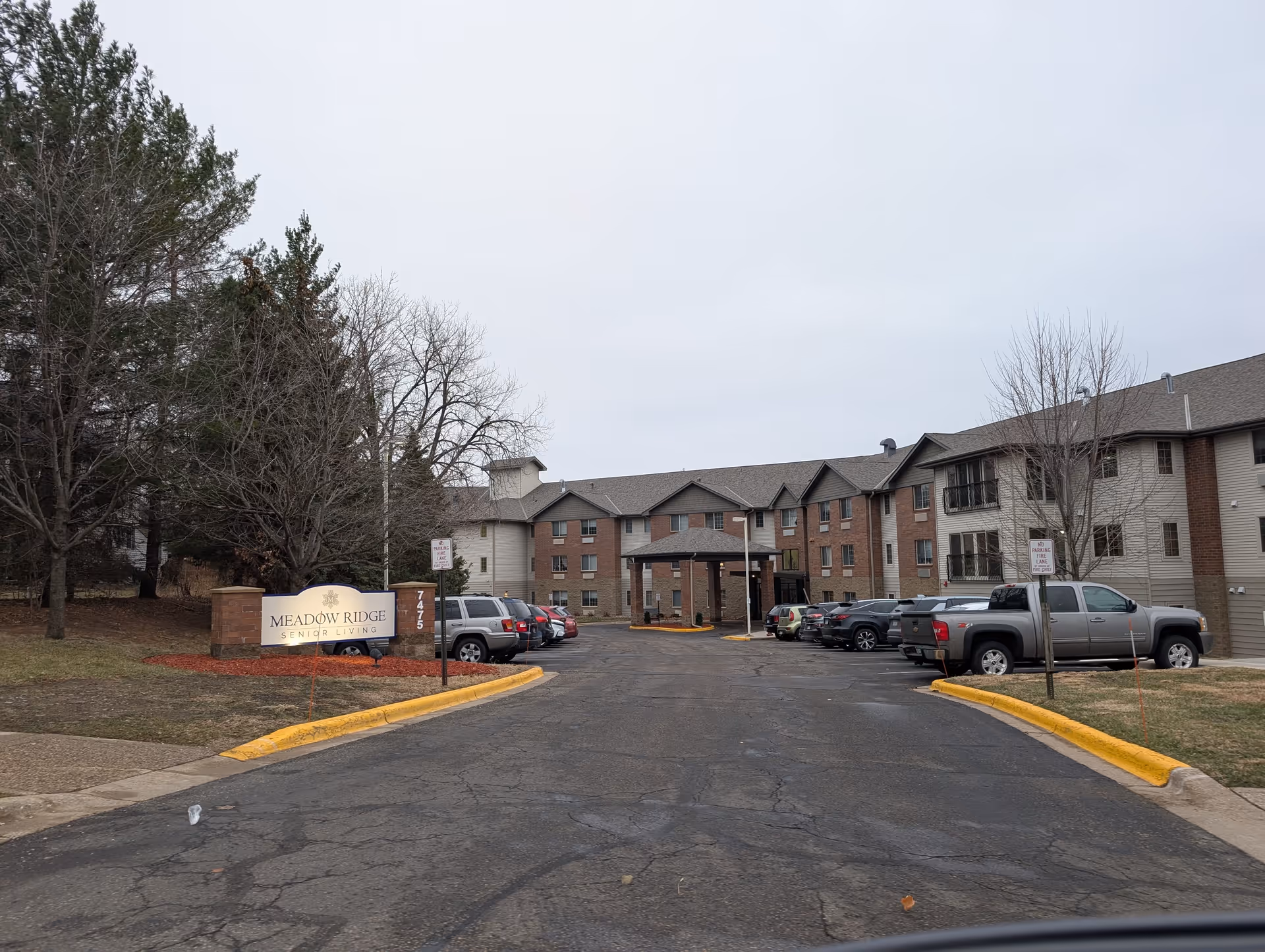 Exterior view of Meadow Ridge Senior Living facility showing a three-story building with a covered entrance, parked cars along the driveway, leafless trees, and a sign with the facility name near the entrance.