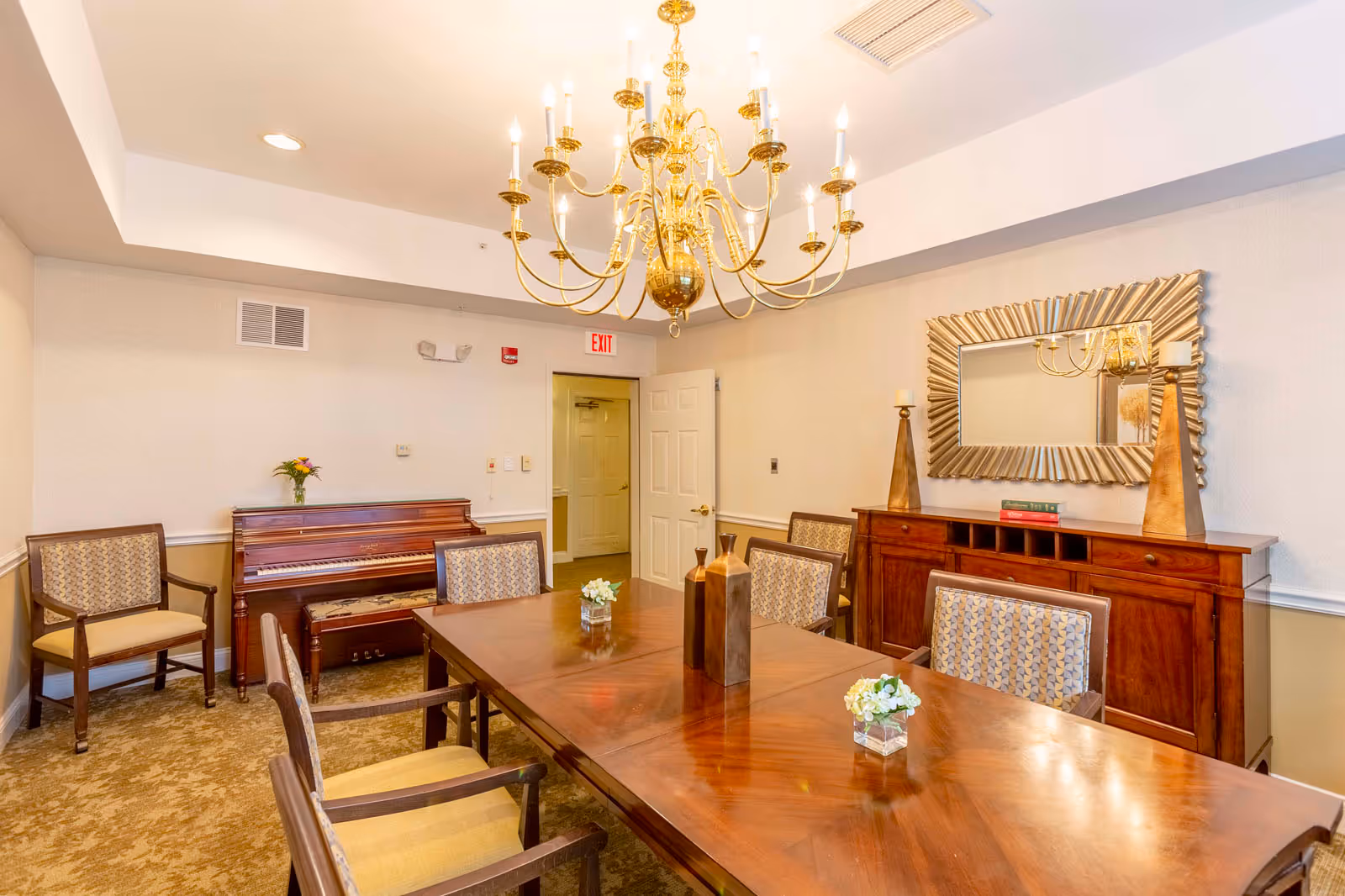 A well-lit dining room with a large wooden table surrounded by upholstered chairs. A decorative gold chandelier hangs from the ceiling. Against one wall is a wooden piano with a small vase of flowers on top, and a large ornate mirror above a wooden sideboard with two tall candle holders and some books. The room has beige walls and carpeted floor, with an open door leading to another area.