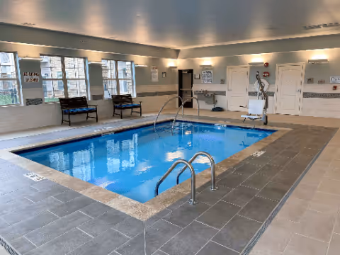 Indoor swimming pool area with clear blue water, surrounded by gray tiled flooring. There are metal handrails for entering the pool, benches along the wall with large windows letting in natural light, and a pool lift for accessibility. The walls are light-colored with some safety and informational signs posted.