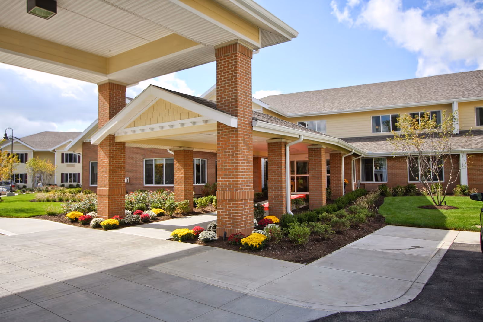 Exterior view of Amber Park Senior Living facility showing a covered entrance with brick pillars, landscaped flower beds with colorful flowers, green lawns, and a clear blue sky with some clouds.