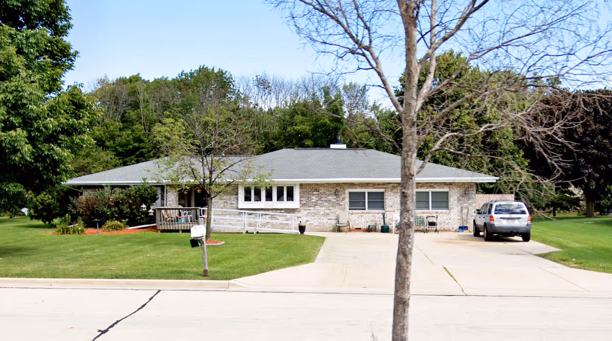 Single-story brick building with a gray roof, a driveway with a parked silver SUV, a small porch with a wheelchair ramp, and a green lawn with trees in front. The background shows dense green trees under a clear blue sky.
