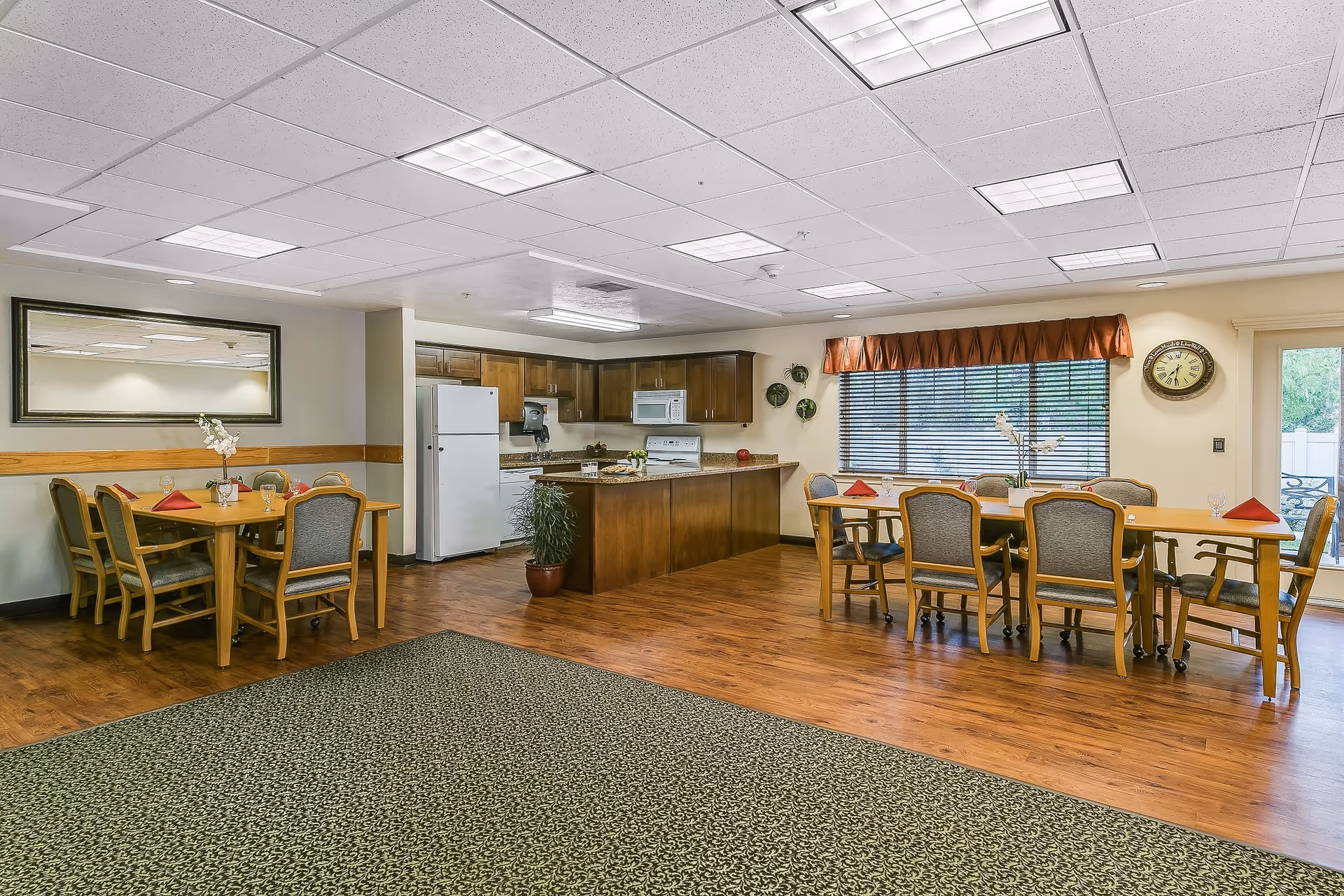 Interior view of a senior living facility dining area with two wooden dining tables, each surrounded by chairs. The tables are set with red napkins and glassware. In the background, there is a kitchen area with wooden cabinets, a white refrigerator, microwave, and stove. A large window with blinds and a brown valance lets in natural light. A wall clock is mounted near the window, and a large mirror hangs on the opposite wall. The floor is a combination of wood and patterned carpet.