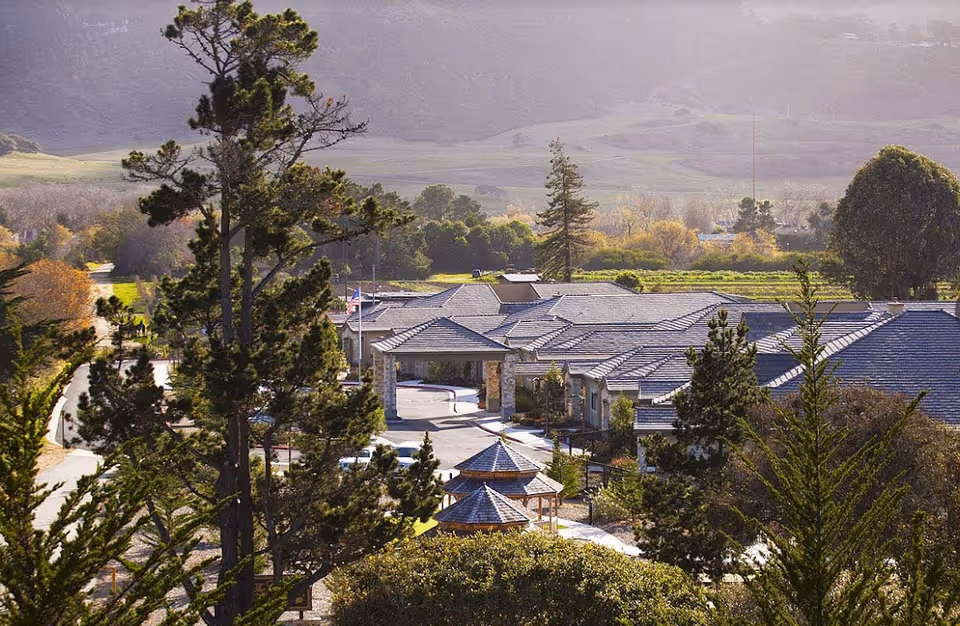 Aerial view of The Cottages Of Carmel senior living facility surrounded by trees and greenery with mountains in the background under a hazy sky.