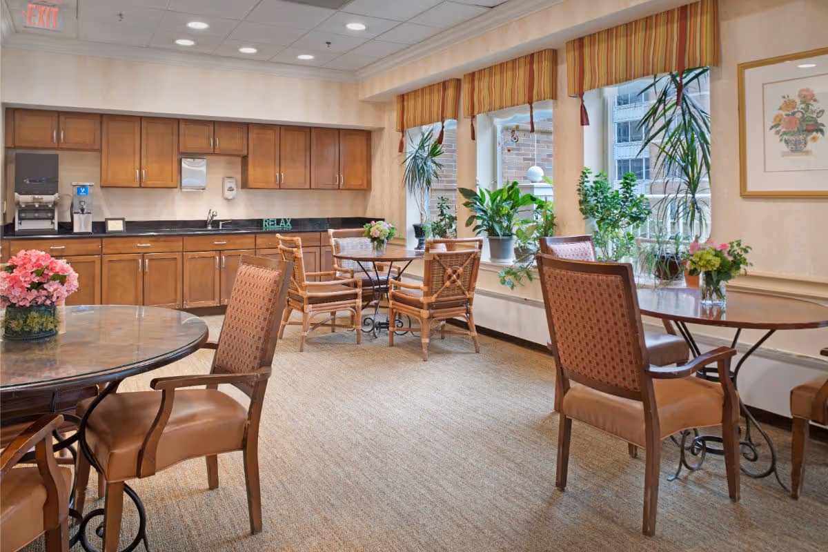 A cozy common area with several tables and chairs arranged for seating. The room features a kitchenette with wooden cabinets, a coffee machine, and a sink. Large windows with striped valances let in natural light and have several potted plants on the windowsill. The room is carpeted and decorated with floral arrangements and a framed picture on the wall.