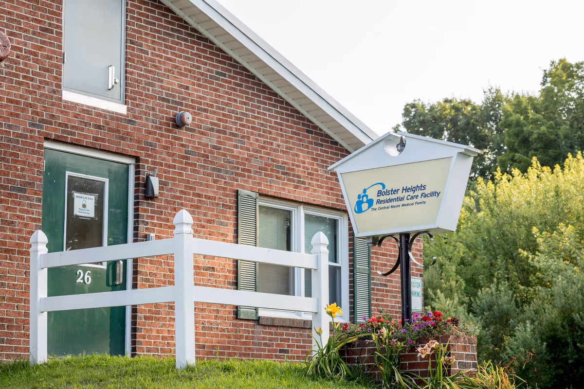 Exterior view of Bolster Heights Residential Care facility showing a brick building with a green door numbered 26, a white fence, a sign with the facility's name, and some flowers and greenery in the foreground.