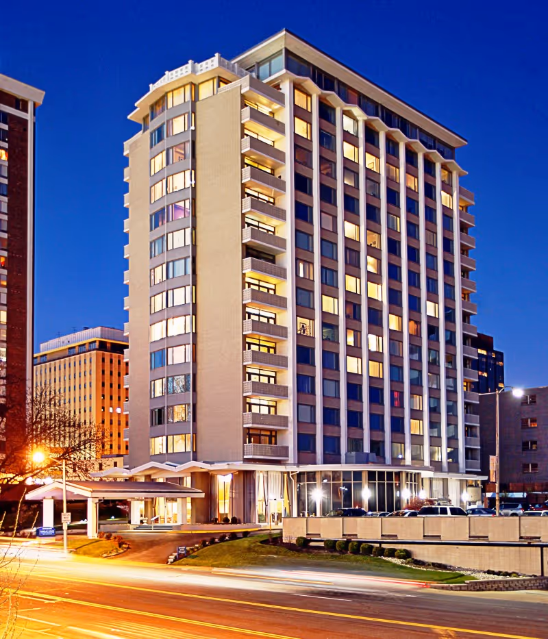 Exterior view of a multi-story senior living facility building named Bethesda Barclay House at dusk with lights on in many windows and cars parked outside.