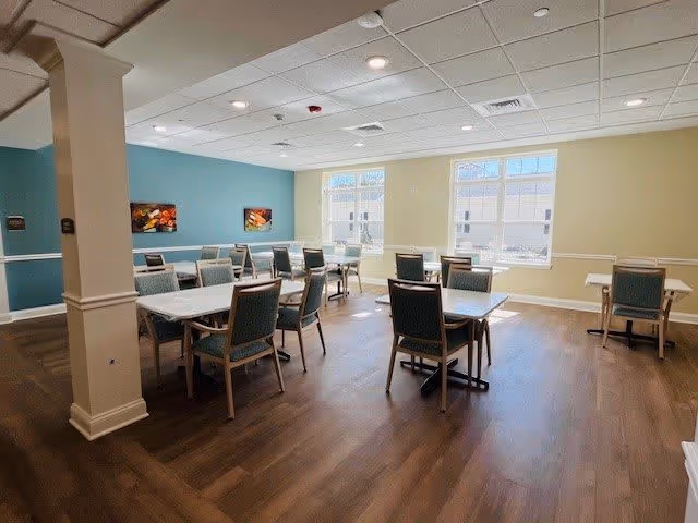 A bright dining room with several tables and chairs arranged neatly on a wooden floor. The walls are painted in light blue and beige tones, with two large windows letting in natural light. There are two colorful paintings on the blue wall and a white column in the foreground.