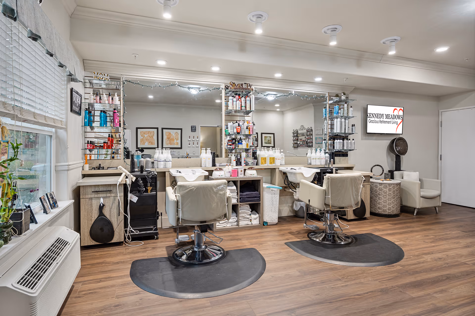 Interior view of a hair salon area in a senior living facility with two salon chairs in front of a large mirror, shelves stocked with hair care products, and a hair dryer chair in the corner. The room has wood flooring, bright lighting, and a window with blinds on the left side. A sign on the wall reads 'Kennedy Meadows Gracious Retirement Living.'