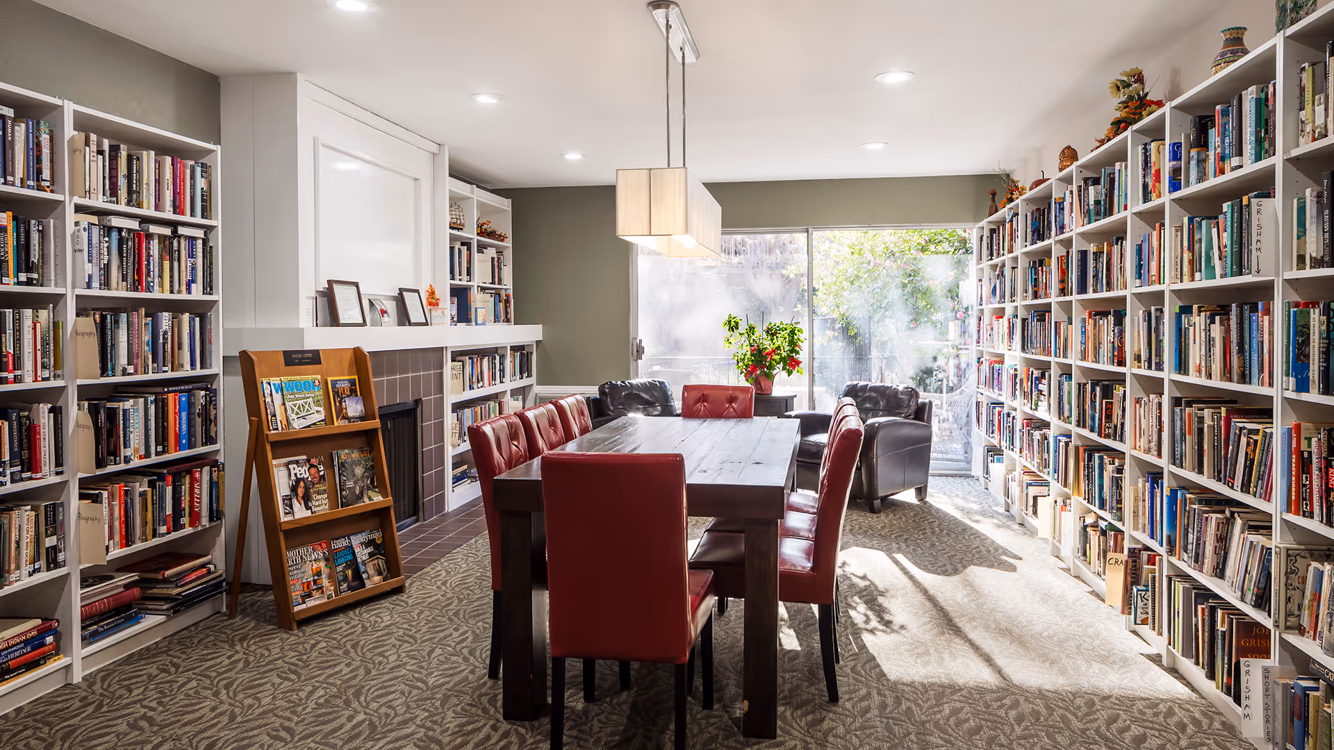 A bright and cozy library room with tall white bookshelves filled with books lining both sides. In the center, there is a dark wooden table surrounded by red leather chairs. At the far end, two black armchairs are positioned near a large window letting in natural light. A small wooden magazine rack stands near a white fireplace on the left side.