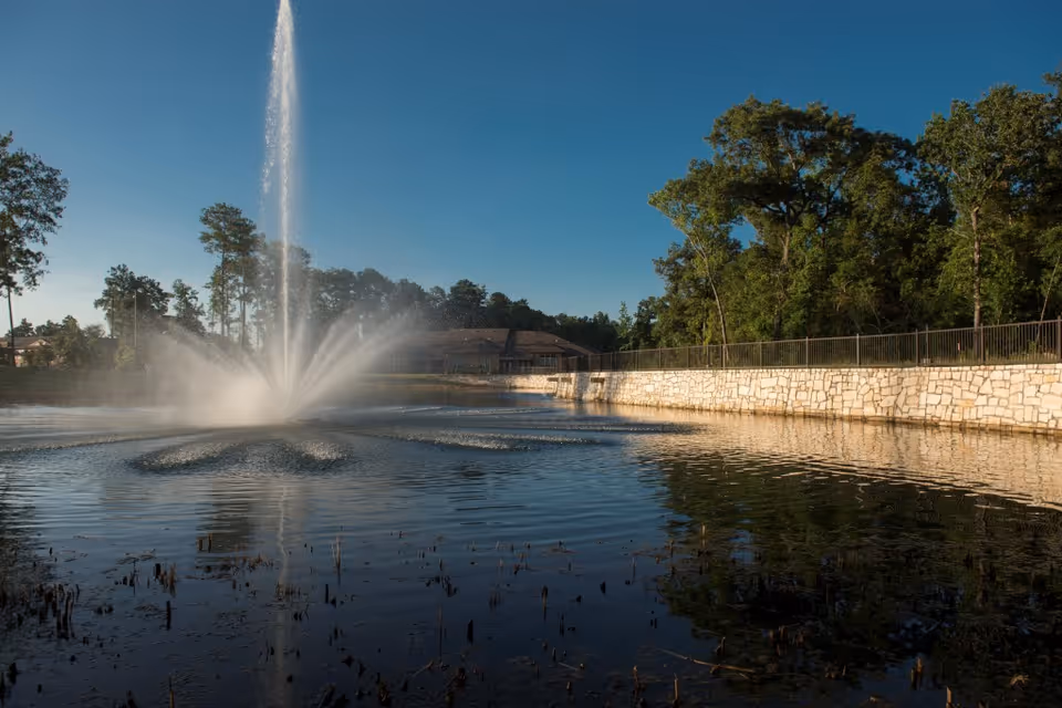 A large water fountain spraying water high into the air in the middle of a pond, surrounded by trees and a stone wall with a metal fence on top under a clear blue sky.